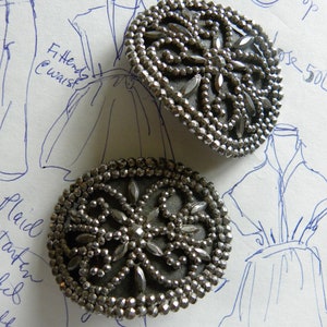 May include: Two antique silver-toned buttons with intricate beaded and faceted designs. The buttons have a central floral pattern and are set against a background of handwritten fashion sketches.