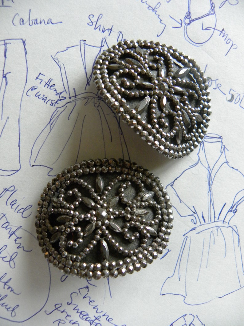 May include: Two antique silver-toned buttons with intricate beaded and faceted designs. The buttons have a central floral pattern and are set against a background of handwritten fashion sketches.