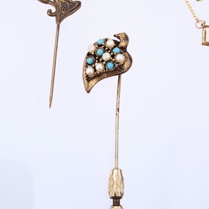 May include: A gold-toned leaf-shaped brooch with white and turquoise blue stones. The brooch is attached to a long, thin pin.