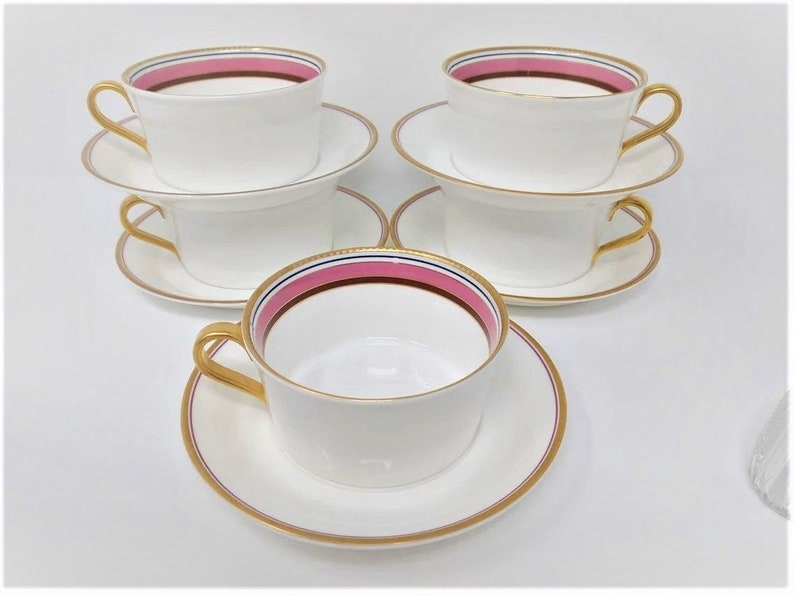 May include: Set of four white teacups and saucers with gold trim and pink and blue stripes around the rim.