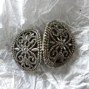 May include: Two silver-toned, oval-shaped clasps with intricate floral and beaded designs. The clasps are open, revealing their inner workings. The background is a crumpled white paper, providing a neutral backdrop for the detailed metalwork.
