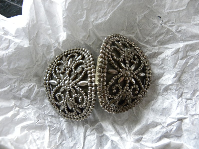 May include: Two silver-toned, oval-shaped clasps with intricate floral and beaded designs. The clasps are open, revealing their inner workings. The background is a crumpled white paper, providing a neutral backdrop for the detailed metalwork.