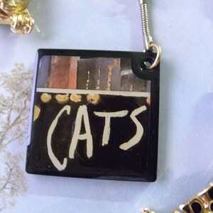 May include: A black and white keychain with a picture of a stage and the word "CATS" written in white paint.