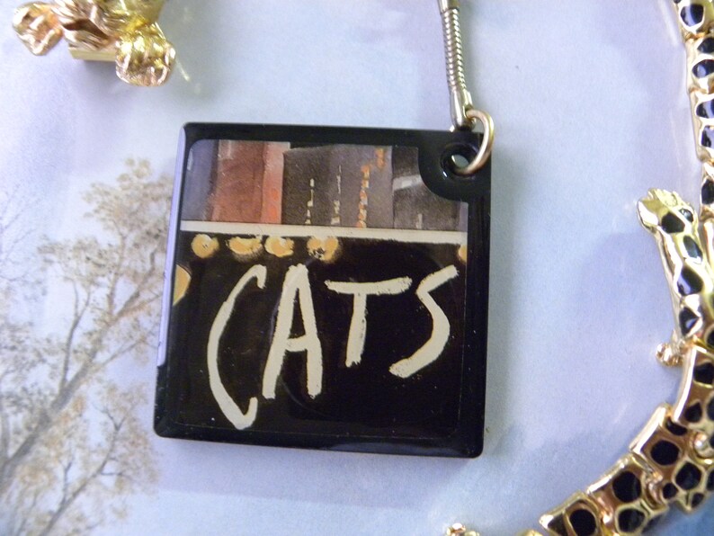 May include: A black and white keychain with a picture of a stage and the word "CATS" written in white paint.