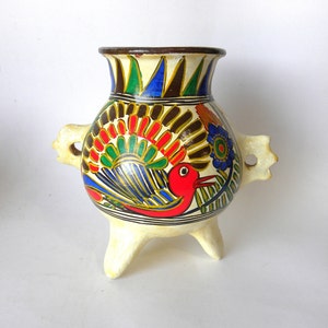 May include: A white ceramic pot with three legs and two handles. The pot is decorated with colorful painted designs of birds, flowers, and geometric patterns.