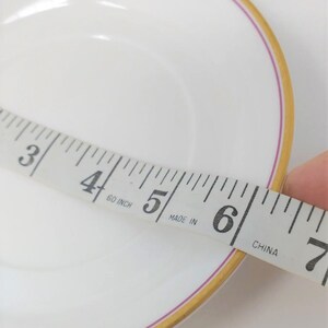 May include: A white saucer with a gold and pink trim. The saucer is being measured with a tape measure that reads "60 inch" and "Made in China".