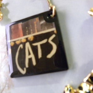 May include: A black and gold necklace with a pendant that says "CATS" in white lettering. The pendant is a rectangular piece of black plastic with a picture of a bookshelf on it.