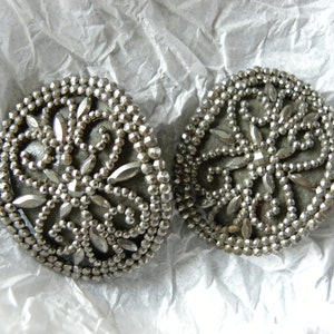 May include: Two silver-toned, round vintage buttons with intricate beaded and faceted detailing. The buttons feature a central floral design with radiating petal shapes. They rest on a crumpled white paper surface.