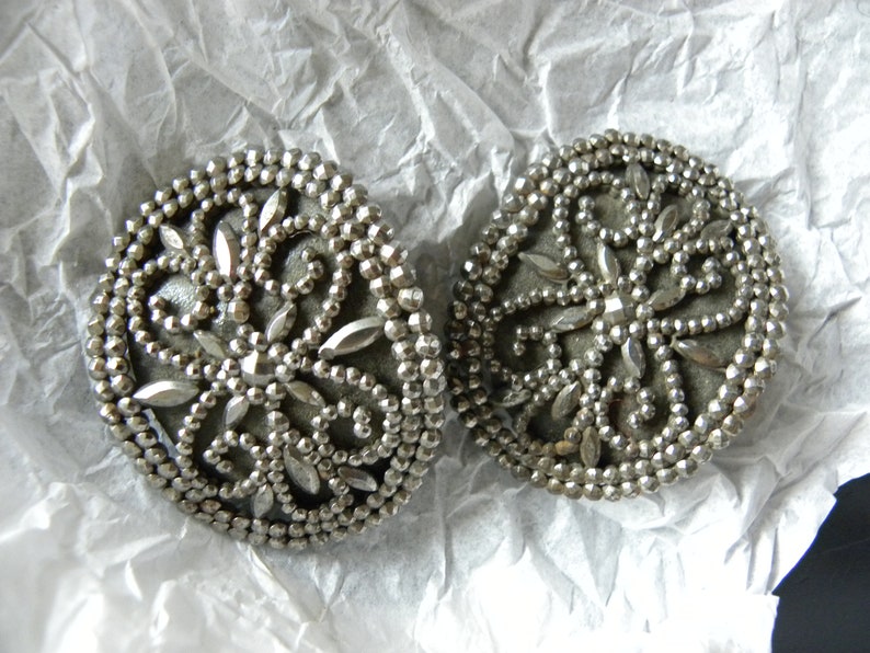 May include: Two silver-toned, round vintage buttons with intricate beaded and faceted detailing. The buttons feature a central floral design with radiating petal shapes. They rest on a crumpled white paper surface.