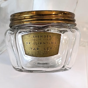 May include: A clear glass jar with a gold-colored metal lid. The jar has a gold label with the text "LAVENDER SOLID FIED BRILLIANTINE YARDLEY LONDON ENGLAND". The jar is sitting on a white surface.