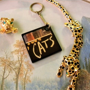 May include: A gold-tone keychain with a black square charm featuring the word "CATS" in white lettering. The charm has a colorful image of a stage set in the background. The keychain is accompanied by a gold-tone tiger pin with green eyes and a gold-tone leopard necklace with black spots.