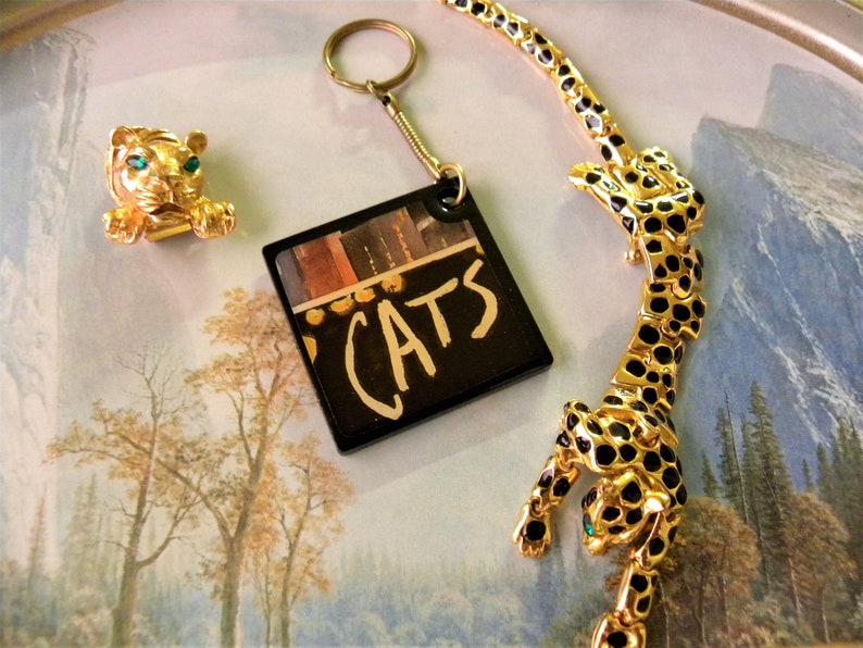 May include: A gold-tone keychain with a black square charm featuring the word "CATS" in white lettering. The charm has a colorful image of a stage set in the background. The keychain is accompanied by a gold-tone tiger pin with green eyes and a gold-tone leopard necklace with black spots.