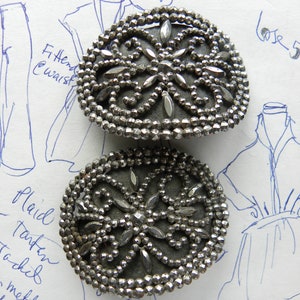 May include: Two antique silver-toned metal buttons. Each button has a dark gray center with a decorative floral design and is surrounded by small, faceted beads. The buttons are on a paper background with handwritten notes and sketches.