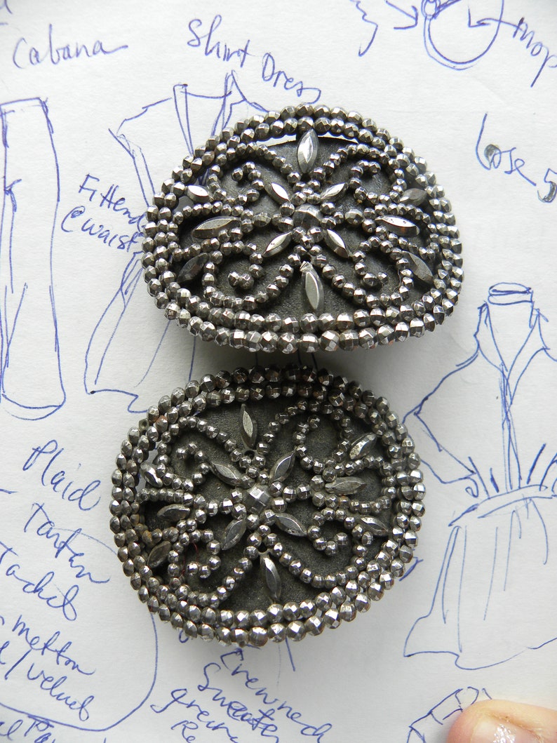 May include: Two antique silver-toned metal buttons. Each button has a dark gray center with a decorative floral design and is surrounded by small, faceted beads. The buttons are on a paper background with handwritten notes and sketches.