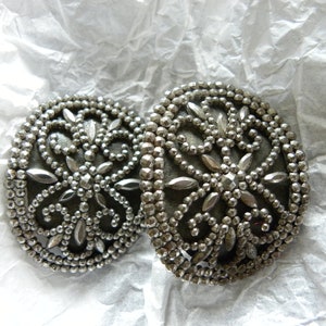May include: Two silver-toned, round buttons with intricate floral designs. The buttons feature a beaded border and a central pattern of stylized leaves and petals. The buttons are displayed on a crumpled white paper surface.