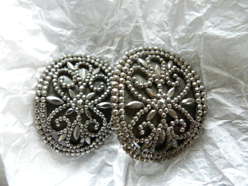 May include: Two silver-toned, round buttons with intricate floral designs. The buttons feature a beaded border and a central pattern of stylized leaves and petals. The buttons are displayed on a crumpled white paper surface.