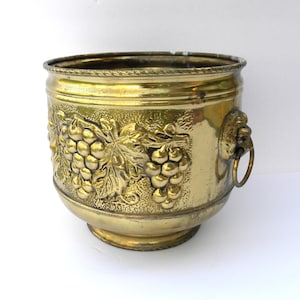 May include: A vintage brass planter with a detailed grape and leaf design. The planter has a round shape with a decorative lion's head handle. The brass has a warm, golden hue, and the planter is likely used for decorative purposes.
