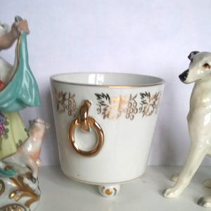 May include: A white porcelain vessel featuring a gold-toned grape vine pattern around the rim, two gold-toned ring handles on the sides, and a single central footed base with gold accents, positioned between two decorative ceramic figurines.