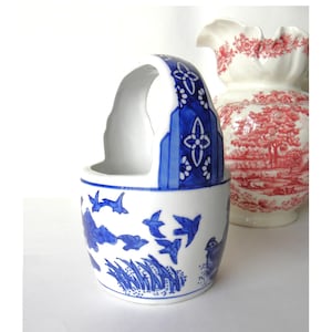 May include: A white ceramic basket with a blue handle and bird and floral designs. The basket has a curved handle and a round base. A red and white floral vase is in the background.