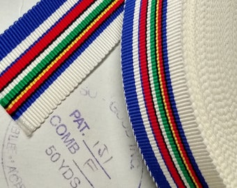 May include: Close-up of a white ribbon with blue, red, green, and yellow stripes. The ribbon is folded and coiled, with a section laid flat. The text "COMB. F 50 YDS." and "MADE IN USA" is visible.