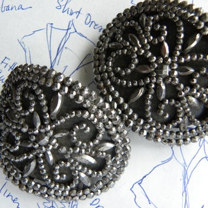 May include: Two antique silver-toned metal buttons with intricate beaded and faceted detailing. The buttons have a dark gray backing and a floral design. The buttons are round and are likely for clothing or decorative purposes.