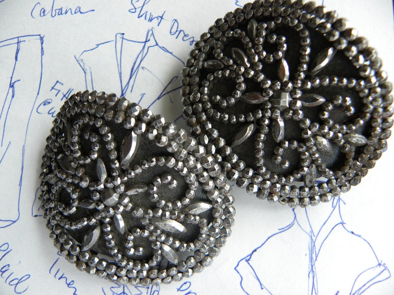 May include: Two antique silver-toned metal buttons with intricate beaded and faceted detailing. The buttons have a dark gray backing and a floral design. The buttons are round and are likely for clothing or decorative purposes.
