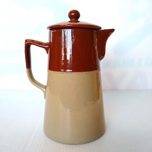 May include: A brown and beige ceramic coffee pot with a lid and a handle. The pot has a rounded body and a spout.