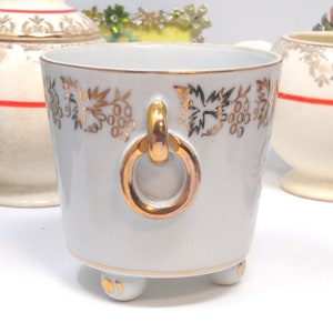 May include: White ceramic planter with a gold ring handle and gold grapevine design. The planter has three gold feet.