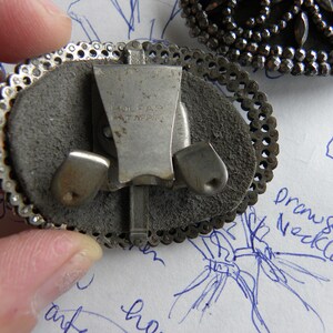 May include: Close-up of a vintage silver-toned clip-on earring with an oval shape. The earring features a decorative filigree border and a gray suede-like backing. The clip mechanism is visible, with the text "HOLFAR PAT. APPL." stamped on it. Another earring is partially visible.