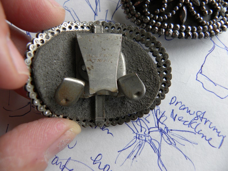 May include: Close-up of a vintage silver-toned clip-on earring with an oval shape. The earring features a decorative filigree border and a gray suede-like backing. The clip mechanism is visible, with the text "HOLFAR PAT. APPL." stamped on it. Another earring is partially visible.
