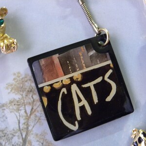 May include: A black square keychain with a colorful image and the word "CATS" written in white paint.