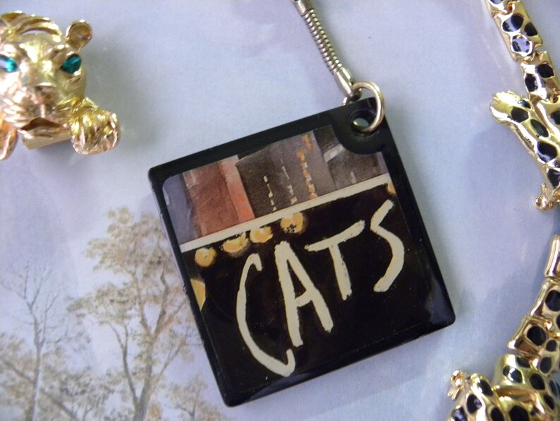 May include: A black square keychain with a colorful image and the word "CATS" written in white paint.