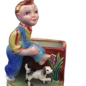 May include: A ceramic planter shaped like a boy in blue overalls kicking a small white dog. The planter has a brown rectangular base with green and pink flowers.