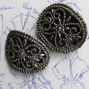 May include: Two ornate, silver-toned metal buttons with intricate beaded designs. The buttons are circular with a floral pattern and are set against a background of fashion sketches and handwritten notes.