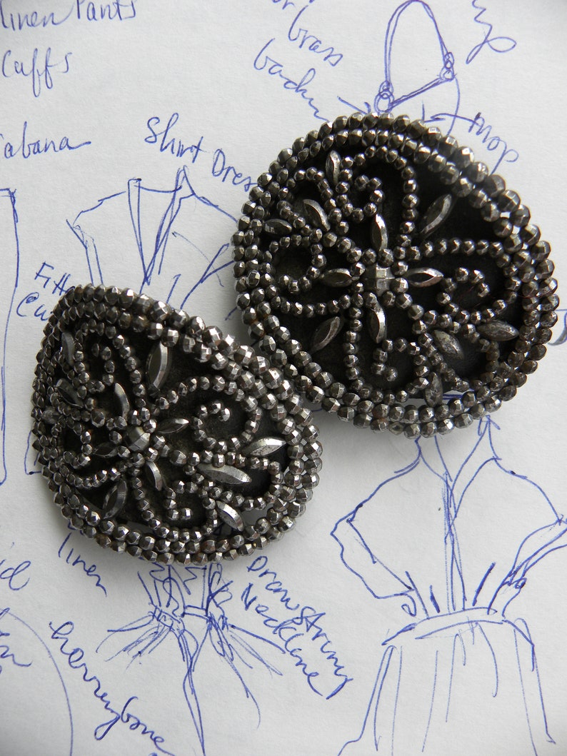 May include: Two ornate, silver-toned metal buttons with intricate beaded designs. The buttons are circular with a floral pattern and are set against a background of fashion sketches and handwritten notes.