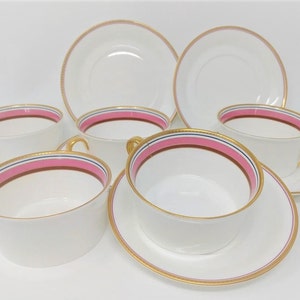 May include: Set of six white porcelain teacups and saucers with pink and gold trim. The cups have a wide, shallow bowl and a handle. The saucers have a raised rim.