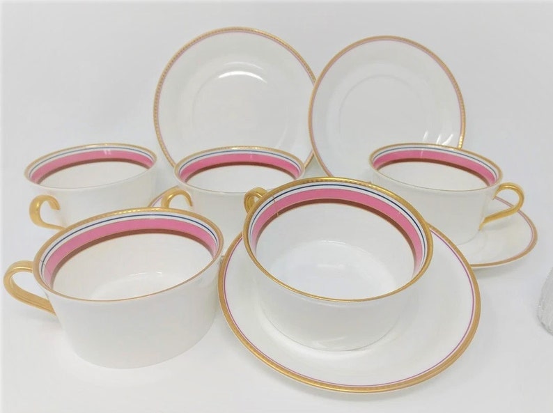 May include: Set of six white porcelain teacups and saucers with pink and gold trim. The cups have a wide, shallow bowl and a handle. The saucers have a raised rim.