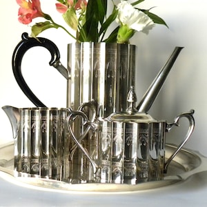 May include: A silver tea set with a black handle on the teapot, a creamer, a sugar bowl, and a tray. The set is decorated with an embossed design.