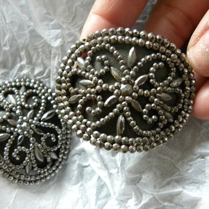 May include: Two antique silver-toned buttons with intricate floral designs. Each button features a central dark gray disc surrounded by a detailed pattern of small beads and leaf-shaped accents. The buttons are round and approximately 1.5 inches in diameter.