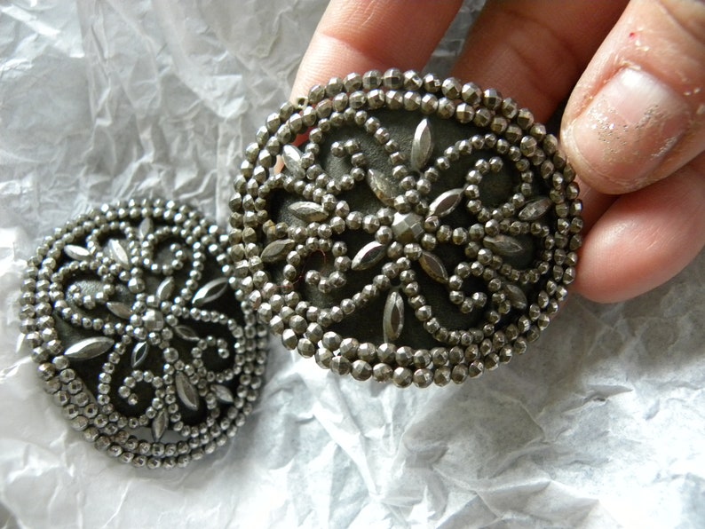 May include: Two antique silver-toned buttons with intricate floral designs. Each button features a central dark gray disc surrounded by a detailed pattern of small beads and leaf-shaped accents. The buttons are round and approximately 1.5 inches in diameter.
