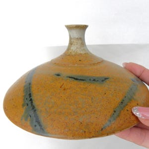 May include: A handmade ceramic vase with a wide, rounded body and a narrow neck. The vase has a speckled, orange-brown glaze with dark gray-blue streaks. The vase is held by a hand.
