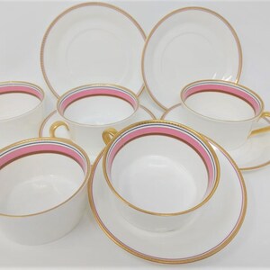 May include: Set of six white porcelain teacups and saucers with pink and gold trim. The cups and saucers have a delicate gold rim and a pink band with a thin gold line.