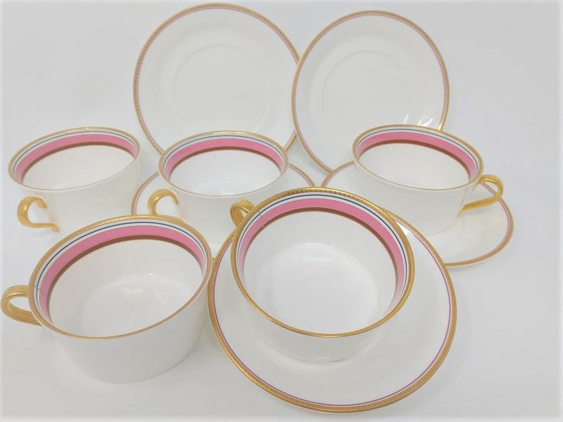 May include: Set of six white porcelain teacups and saucers with pink and gold trim. The cups and saucers have a delicate gold rim and a pink band with a thin gold line.