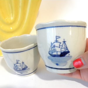 May include: Two white ceramic egg cups with blue and white sailboat designs. The egg cups have a scalloped rim.