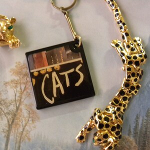May include: A gold-tone keychain with a black square charm that says "CATS" in white lettering. There is also a gold-tone cat brooch with green eyes and a gold-tone necklace with a black and gold leopard design.