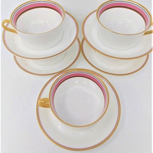 May include: Set of three white teacups and saucers with pink and gold trim. The cups and saucers have a delicate gold rim and a pink band with a thin blue stripe.