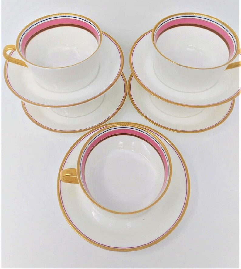 May include: Set of three white teacups and saucers with pink and gold trim. The cups and saucers have a delicate gold rim and a pink band with a thin blue stripe.