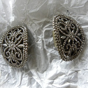 May include: Two silver-toned, half-moon shaped vintage earrings. Each earring features a detailed floral design with small, round beads and elongated, petal-shaped accents. The earrings are displayed on a crumpled white surface.