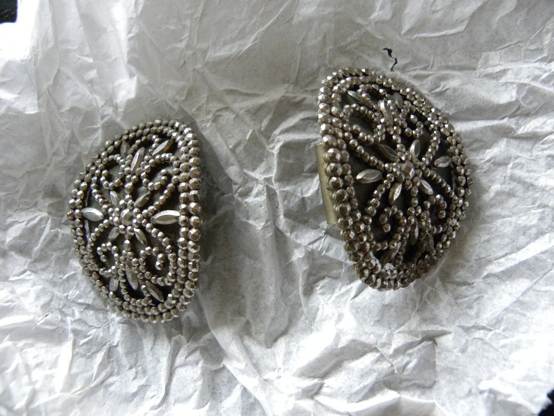May include: Two silver-toned, half-moon shaped vintage earrings. Each earring features a detailed floral design with small, round beads and elongated, petal-shaped accents. The earrings are displayed on a crumpled white surface.