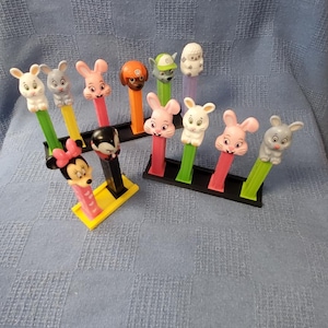 Può includere: Una collezione di dieci dispenser Pez colorati con personaggi, tra cui Topolino, un coniglio, un cane e una pecora. I dispenser sono esposti su stand neri e gialli.